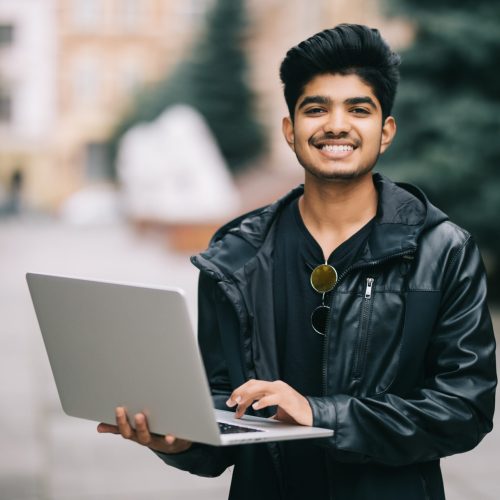 Young indian man standing outdoor with laptop in front of business building